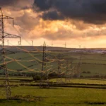 High-voltage electricity pylons and wind turbines across the British countryside at dusk, illustrating the national grid