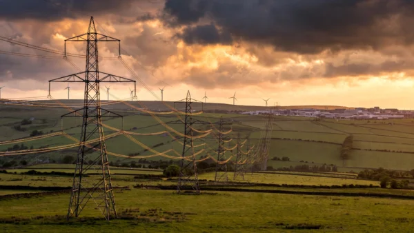 High-voltage electricity pylons and wind turbines across the British countryside at dusk, illustrating the national grid