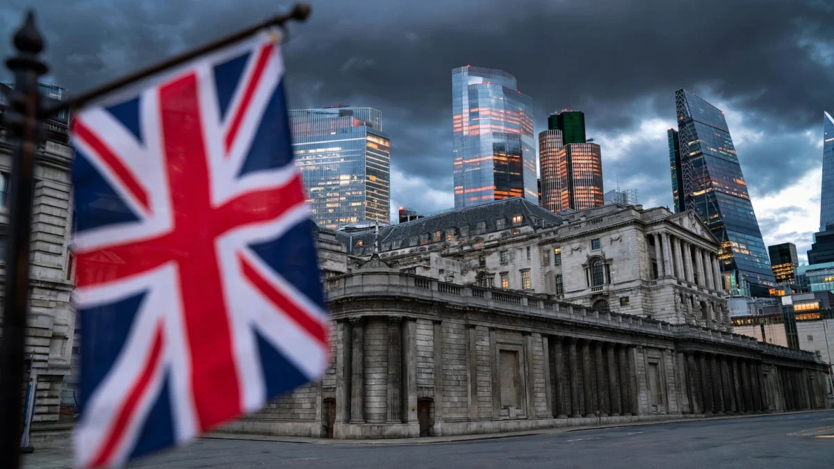 Bank of England building in Threadneedle Street, London, with a Union Jack flag and City of London skyscrapers under a dark dusk sky