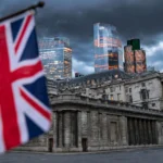 Bank of England building in Threadneedle Street, London, with a Union Jack flag and City of London skyscrapers under a dark dusk sky