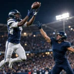 A wide receiver in navy and white leaps for a catch under bright stadium floodlights at night as a defender closes in
