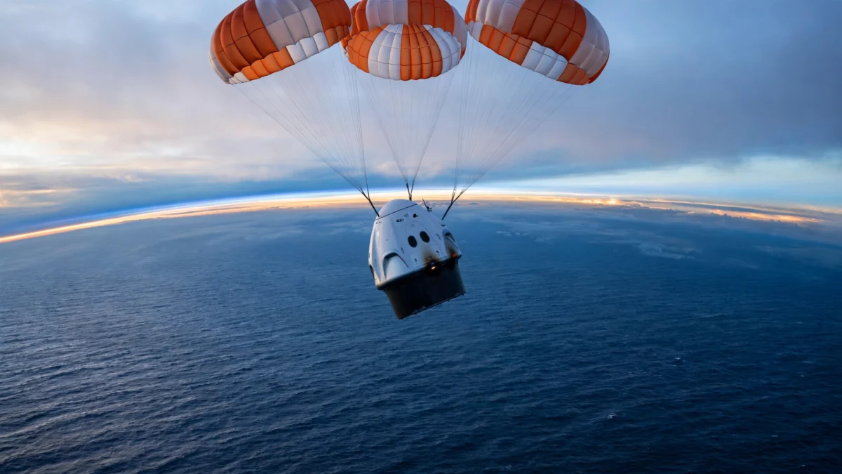 A SpaceX Dragon capsule descending under three orange-and-white parachutes toward the Pacific Ocean at dawn, with the Earth's curved horizon glowing behind it.