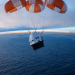 A SpaceX Dragon capsule descending under three orange-and-white parachutes toward the Pacific Ocean at dawn, with the Earth's curved horizon glowing behind it.