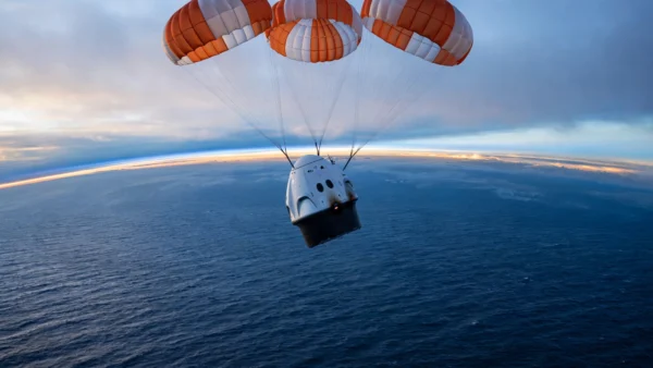 A SpaceX Dragon capsule descending under three orange-and-white parachutes toward the Pacific Ocean at dawn, with the Earth's curved horizon glowing behind it.