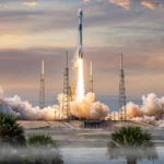 A SpaceX Falcon 9 rocket with a Dragon capsule lifts off from the Cape Canaveral launch pad at dawn, trailing flame and exhaust clouds over palm trees.