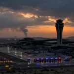 Modern Middle Eastern international airport at dusk with control tower, thin smoke column on the horizon and emergency vehicles on the tarmac