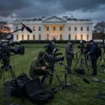 Television news camera crews with broadcast cameras set up on the White House lawn at dusk.