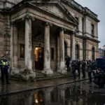Exterior of Westminster Magistrates' Court in London with police officers and press photographers on the steps at dusk.