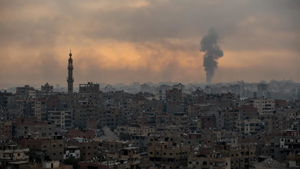 Gaza City skyline at dusk with a distant column of smoke rising over rooftops, illustrating Israeli strikes on the first anniversary of the Gaza ceasefire
