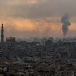 Gaza City skyline at dusk with a distant column of smoke rising over rooftops, illustrating Israeli strikes on the first anniversary of the Gaza ceasefire