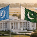 United Nations and Pakistan flags flying side by side outside a government building in Islamabad, with an arriving aircraft in the background