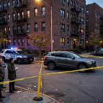 Yellow police tape cordons off a gray SUV on a Bronx residential street at dusk with NYPD patrol cars and onlookers behind the line after an ICE agent shooting in Marble Hill.