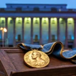 Gold Nobel Prize medal with blue-and-gold ribbon on a wooden lectern before the illuminated Stockholm Concert Hall at dusk.