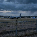 Photorealistic editorial image of a British military airbase at dusk with a large bomber aircraft on the runway and distant emergency service lights, no text.