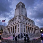 Press photographers on the steps of the federal courthouse in Miami at dusk, with the US flag and police vehicles nearby, after former congressman David Rivera was sentenced to 10 years in prison