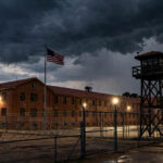 The Texas State Penitentiary at Huntsville at dusk, with a guard tower, barbed-wire fences, floodlights and an American flag under a stormy sky.