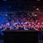 A packed campaign rally arena at night, with a podium on stage, large American flags and supporters waving small flags