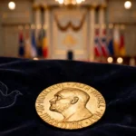 The Nobel Peace Prize medal resting on dark velvet against a softly lit Oslo ceremony hall backdrop, symbolising the 2026 award.