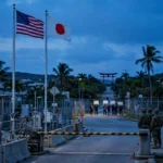 American and Japanese flags fly side by side at dusk outside a US Marine base gate in Okinawa, with a distant crowd holding blank placards and security personnel standing nearby — editorial illustration of the month-long curfew imposed after a Marine's arrest.