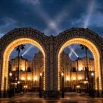 The illuminated front gates of a grand Hollywood film studio at dusk, with searchlights sweeping the evening sky, symbolising the Paramount–Warner Bros merger