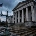 Stone courthouse steps with tall columns under a stormy dusk sky, an American flag flying at left and a bronze lamppost ornament in the foreground, symbolising the reopened Christa Pike lethal-injection case in Tennessee