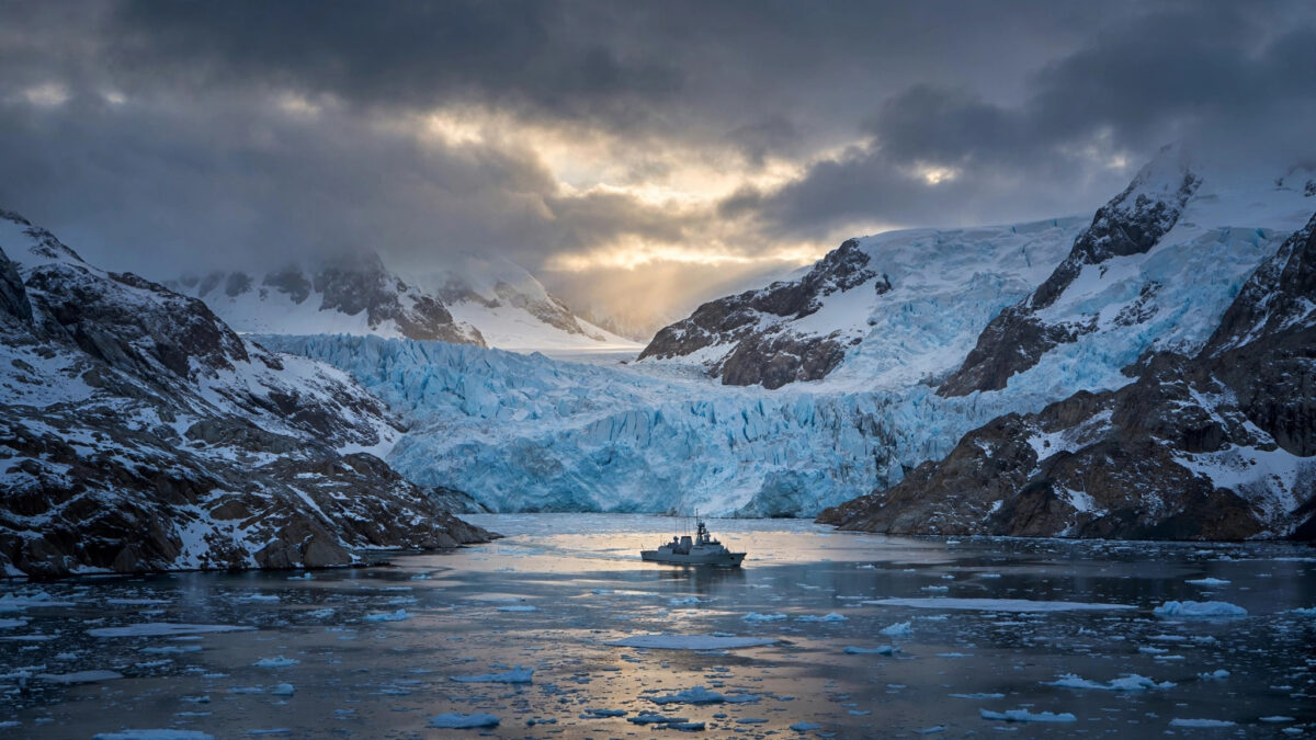A naval vessel crossing an Arctic fjord beneath massive glaciers at dusk