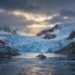 A naval vessel crossing an Arctic fjord beneath massive glaciers at dusk