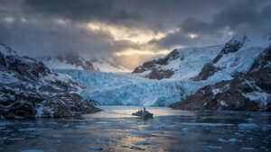 A naval vessel crossing an Arctic fjord beneath massive glaciers at dusk