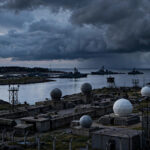 Fortified Russian coastal military installation on the Baltic Sea at dusk, with warships and radar domes under dark storm clouds