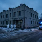 Workers in white hazmat suits at a cordoned-off district hospital in a Siberian town at dusk, with an ambulance flashing lights, during a quarantine after a laboratory worker's death.