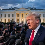 US president speaking to a scrum of reporters outside the White House at dusk, raised microphones, American flag in the background