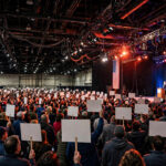 Photorealistic editorial illustration of a packed campaign rally inside a large convention center: a speaker at a podium on stage with large American flags, and a crowd holding blank placards, no text anywhere in the image.