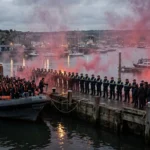 A lifeboat ferrying migrants ashore at a British harbour at dusk, with police forming a cordon as protesters gather and red flares burn