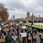 Pro-Palestine demonstrators carrying Palestinian flags and blank placards march along the Thames toward Big Ben and the Houses of Parliament as Metropolitan Police officers in high-visibility jackets line the pavement in central London