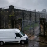 Stone perimeter wall, barbed wire and security gates of a British prison at dawn, with a prison transport van outside the gates