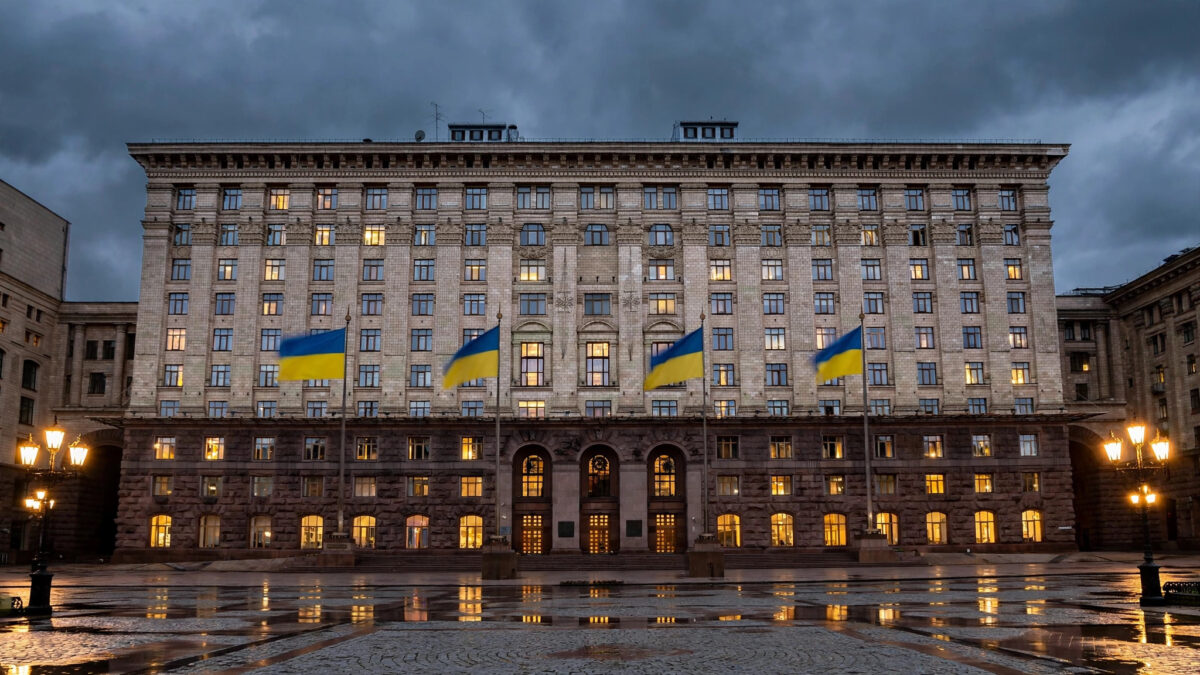 A grand government ministry building in Kyiv at dusk, with large Ukrainian blue-and-yellow flags flying from tall flagpoles in front