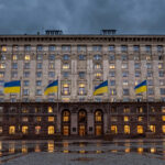 A grand government ministry building in Kyiv at dusk, with large Ukrainian blue-and-yellow flags flying from tall flagpoles in front