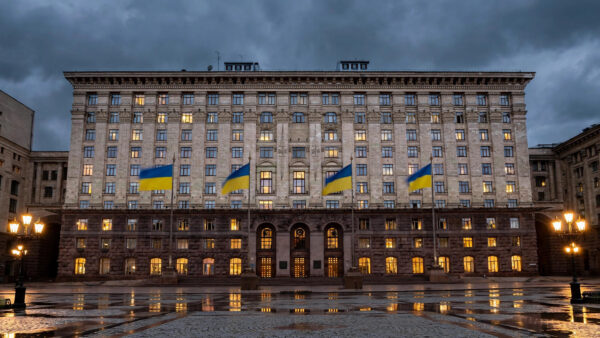 A grand government ministry building in Kyiv at dusk, with large Ukrainian blue-and-yellow flags flying from tall flagpoles in front