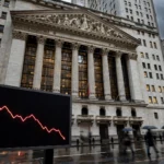 New York Stock Exchange facade at dusk with a large electronic screen showing a red downward-trending stock chart line, rain-wet street