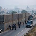 Steel shipping containers stacked as a barricade across a highway into Islamabad, with police personnel and patrol vans beside them and cargo trucks queued on the roadside.
